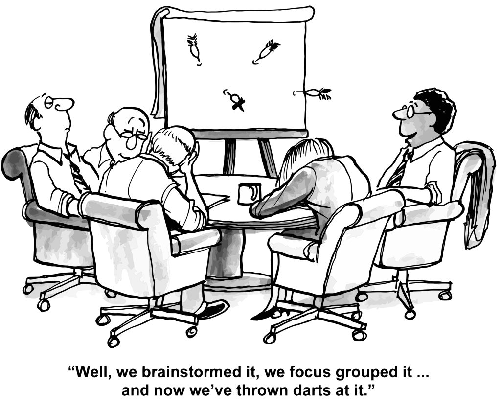 Cartoon showing business executives sitting around a flipchart with darts thrown all over it. The caption reads: "Well, we brainstormed it, we focus grouped it... and now we've thrown darts at it."