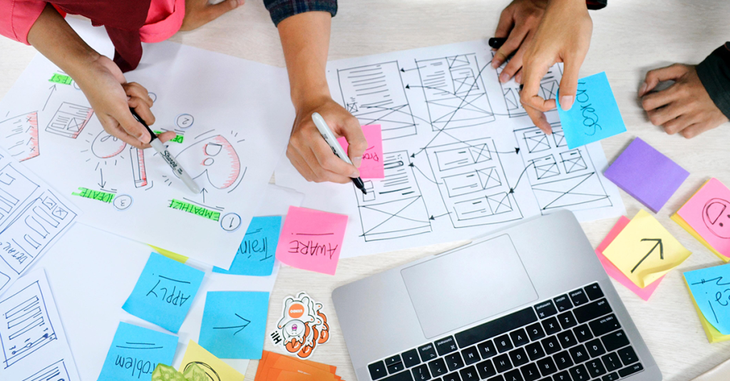 Designers sitting around their design artifacts, such as the design process, their wireframes and wireflows, as well as the outputs from their ideation and ux research.